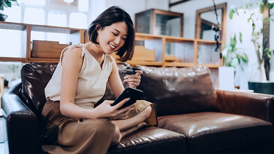 A woman smiling while sitting on a couch looking at her credit card and mobile phone.