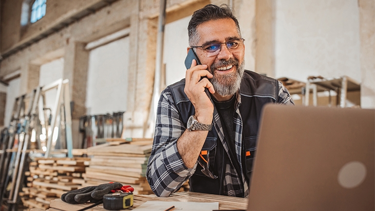 A businessman in his woodworking shop on a mobile phone and using his laptop.