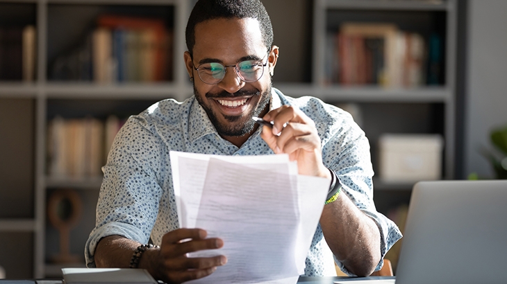 Man smiling and looking at paperwork with laptop.