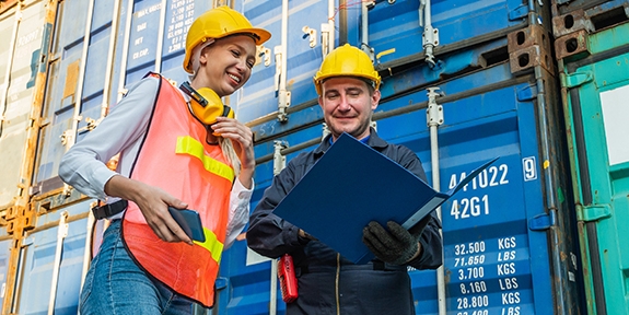 Two people in hardhats talking by shipping containers.