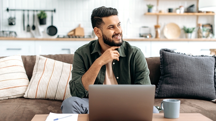 Man sitting at home on sofa using laptop and thinking while smiling.