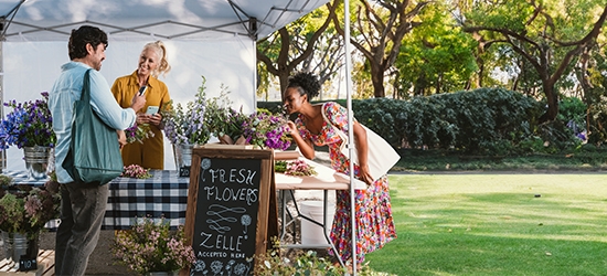 People buying flowers at an outdoor booth with a Zelle accepted here sign.