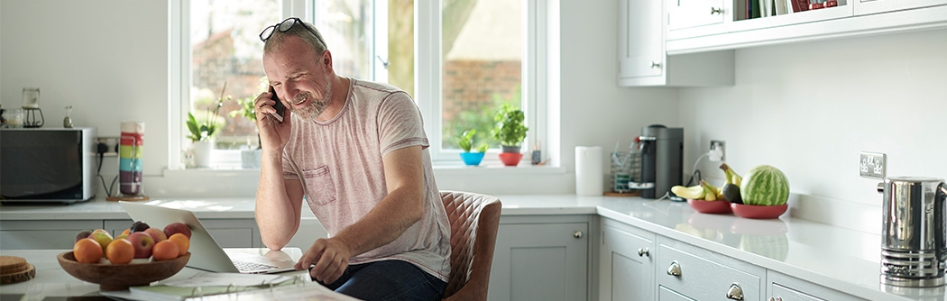 Mature man in home kitchen sorting the home finances on laptop, talking on cell phone