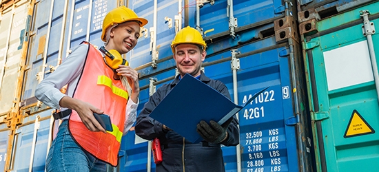 Two people in hardhats talking by shipping containers.